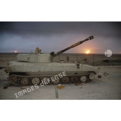 Un canon automoteur irakien de 122 mm M1974 (ou 2S1) abandonné devant un horizon de puits de pétrole en feu.