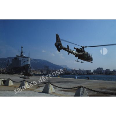 Un hélicoptère de combat Gazelle du 3e RHC (régiment d'hélicoptères de combat) décolle d'un quai du port de Toulon. En arrière-plan le TCD (transport de chalands de débarquement) Foudre est amarré au quai.