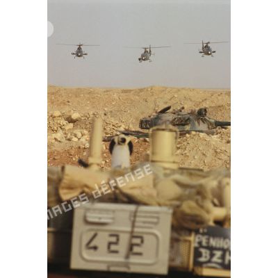 Des hélicoptères de combat Gazelle Hot et Canon volent vers l'objectif avec au premier plan un blindé de reconnaissance AMX-10 RC. Au second plan une tourelle de char de combat T-55 irakien.