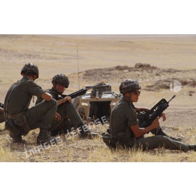 Trois soldats portant le casque lourd et armés de fusil d'assaut FAMAS sont assis au sommet d'une colline dans le désert lors d'un exercice de tir.
