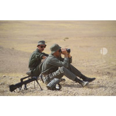 Soldats scrutant le désert à l'aide de jumelles, assis à côté d'un fusil d'assaut FAMAS.