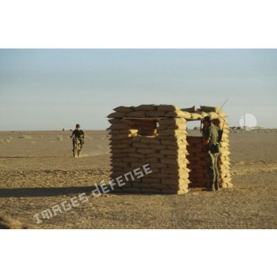 Une sentinelle gardant le camp est postée sous un abri de sacs de sable. Une estafette à moto Peugeot 80 cm3 progresse à proximité.
