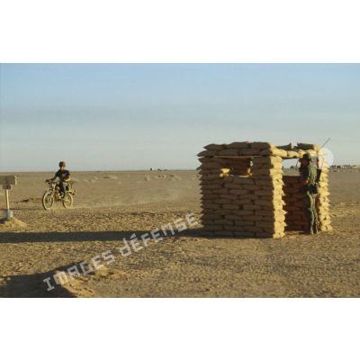 Une sentinelle gardant le camp est postée sous un abri de sacs de sable. Une estafette à moto Peugeot 80 cm3 passe à proximité.