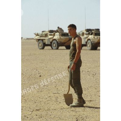 Un soldat est équipé d'une pelle dans le desert, en arrière-plan des VAB.