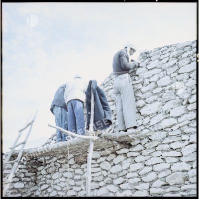 Trois hommes sur un échafaudage le long d’un mur en construction dans la région d’Aïn Tindamine.