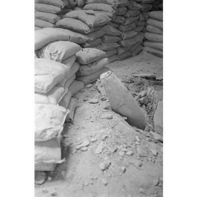 Gros plan sur une bombe alliée qui n'a pas explosé en traversant le toit d'un hangar et en touchant le sol, des sacs de sable sont positionnés autour de l'objectif.