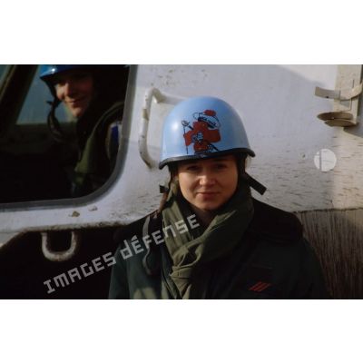 Portrait d'une infirmière du Service de Santé des Armées posant devant un véhicule de l'avant blindé (VAB) et arborant un casque bleu.