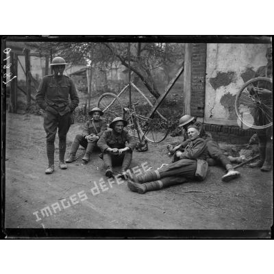 [Des soldats sont assis et allongés sur le bord d'une route de village, pendant que l'un d'entre eux répare la roue d'un vélo.]