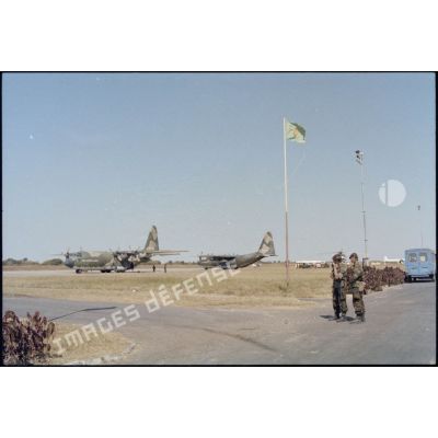 Plan général de l'aéroport de Lubumbashi sous la garde de parachutistes belges. Au dessus des deux militaires flotte le drapeau zaïrois ; à l'arrière-plan, deux avions de transport Hercules C130 zaïrois. [Description en cours]