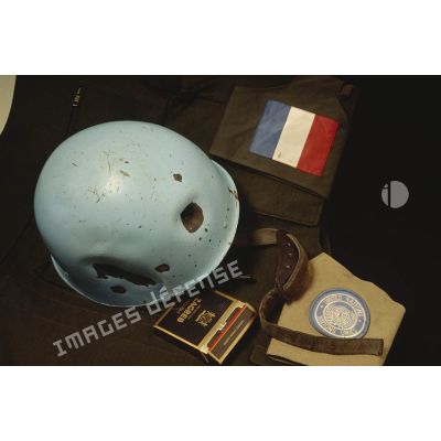 Casque bleu d'un sergent du 8e régiment d'infanterie (8e RI) ayant été blessé à la tête par balle en ex-Yougoslavie posé sur une veste militaire avec le drapeau français, l'insigne de l'ONU et un paquet de cigarettes croates.