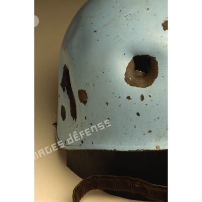 Gros plan sur des impacts sur le côté d'un casque bleu mis en scène sur fond blanc et appartenant à un sergent du 8e régiment d'infanterie (8e RI) ayant été blessé à la tête par balle en ex-Yougoslavie.
