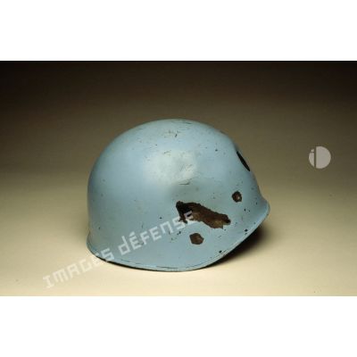 Côté droit d'un casque bleu mis en scène sur fond blanc présentant des impacts et appartenant à un sergent du 8e régiment d'infanterie (8e RI) ayant été blessé à la tête par balle en ex-Yougoslavie.