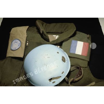 Vue d'au-dessus du casque bleu présentant des impacts d'un sergent du 8e régiment d'infanterie (8e RI) ayant été blessé à la tête par balle en ex-Yougoslavie mis en scène sur un gilet militaire avec le drapeau français et l'insigne de l'ONU.