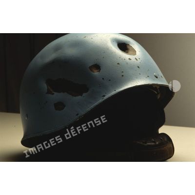 Gros plan sur un casque bleu présentant des impacts mis en scène en clair obscur et appartenant à un sergent du 8e régiment d'infanterie (8e RI) ayant été blessé à la tête par balle en ex-Yougoslavie.