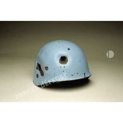 Casque bleu présentant un impact frontal posé sur fond blanc et appartenant à un sergent du 8e régiment d'infanterie (8e RI) ayant été blessé à la tête par balle en ex-Yougoslavie.